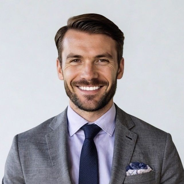 Smiling man in a suit and tie with a pocket square, standing against a light background.