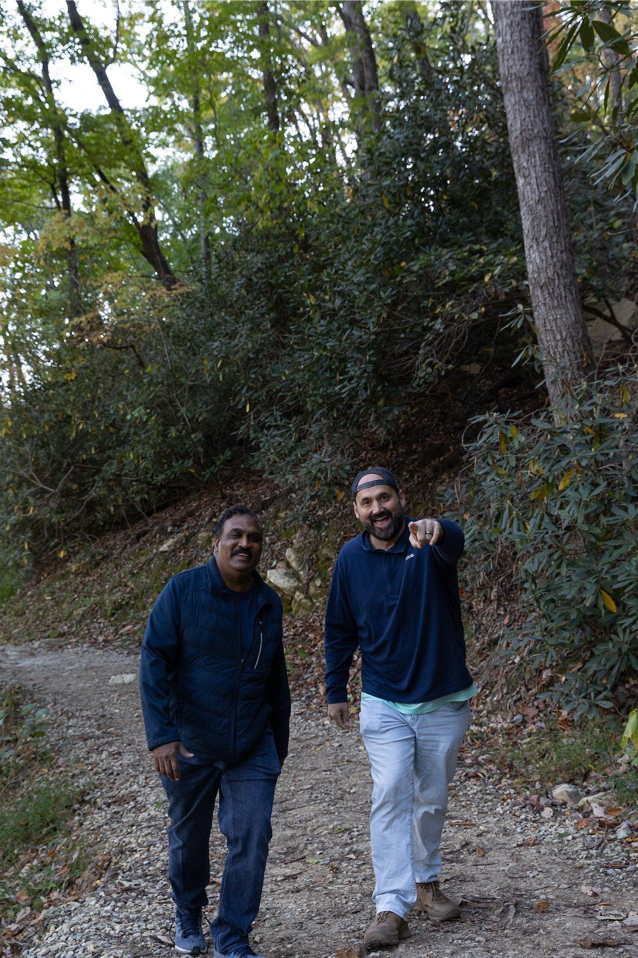 Two individuals walking on a gravel trail surrounded by dense greenery and trees, one person pointing ahead.