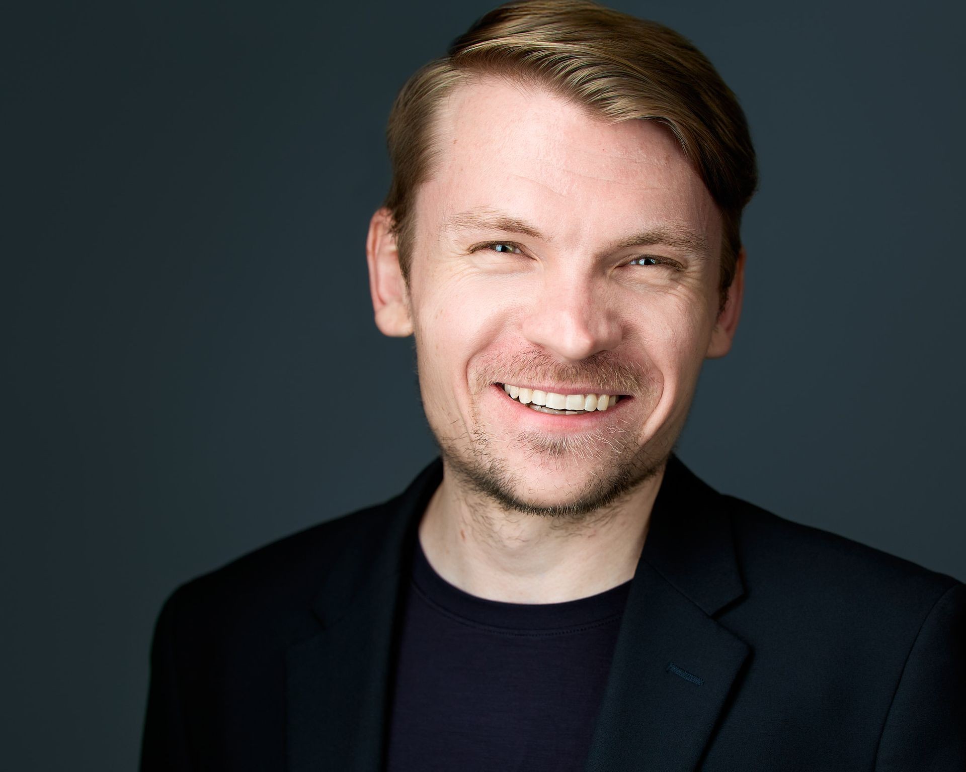 Smiling person with short hair wearing a black blazer against a dark background.