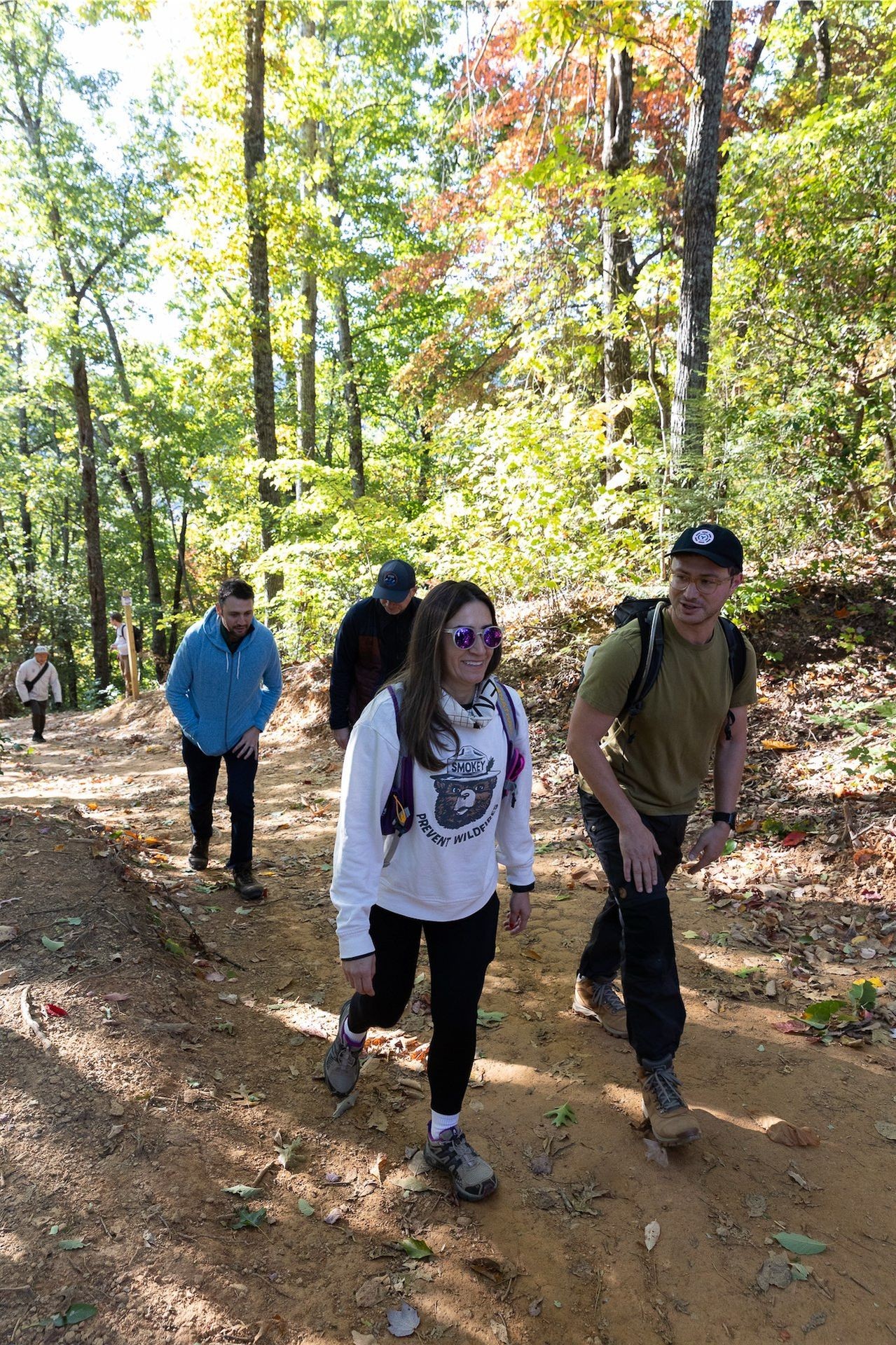 Group of people hiking on a forest trail with autumn foliage, wearing casual outdoor clothing and backpacks.