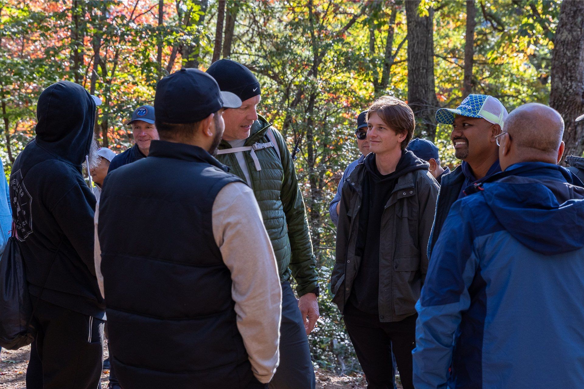 Group of people outdoors wearing jackets and hoodies gathered under sunlight surrounded by trees with autumn leaves.