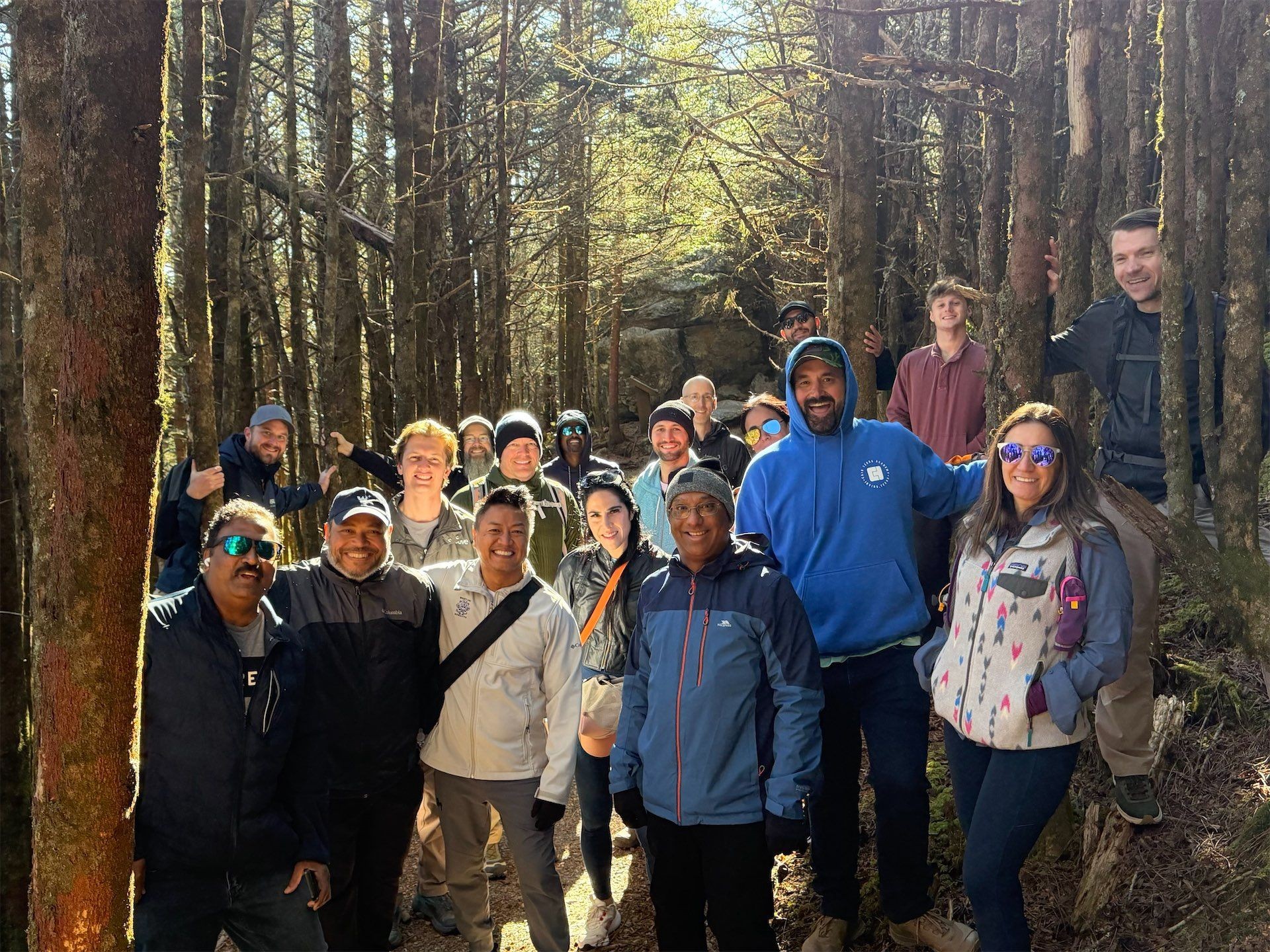Group of people posing together in a sunlit forest with trees in the background.