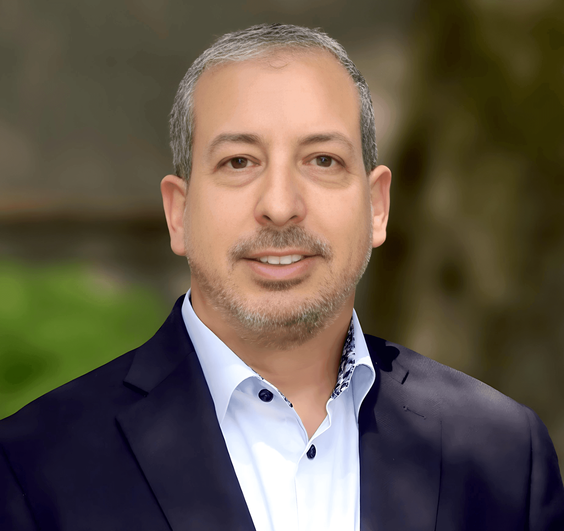 Man in a business suit smiling, standing outdoors with a blurred natural background.